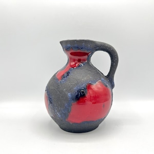 Op de afbeelding: Een kleine, rode en zwarte keramische kan met een getextureerd oppervlak. De kan heeft een breed, rond lichaam en een klein, gebogen handvat.