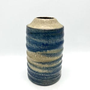 May include: A cylindrical ceramic vase featuring a textured, wavy surface with alternating horizontal bands of deep blue and speckled beige glaze. The vase has a narrow, tapered neck and a wide, open top, showcasing a rustic, hand-crafted appearance.