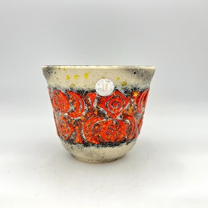 Puede incluir: Una maceta de cerámica con una superficie texturizada y un diseño llamativo. La maceta presenta una base de color crema con un patrón de motivos florales rojos y naranjas. Una pequeña pegatina plateada está adherida al frente.