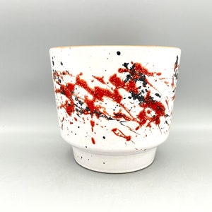 Könnte beinhalten: Weißer Keramiktopf mit einem Spritzmuster in Schwarz und Rot.