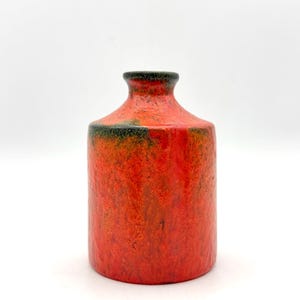 May include: A cylindrical ceramic vase with a vibrant orange and red textured glaze. The surface features mottled dark green and black accents near the narrow neck and rim, creating a distinct contrast against the bright, fiery orange body.