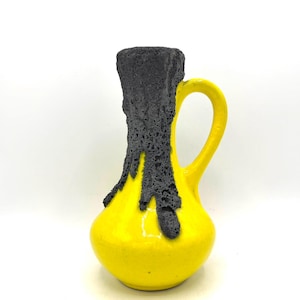 May include: A yellow ceramic pitcher with a black, textured top and handle. The black texture drips down the side of the pitcher. The pitcher has a rounded base and a curved handle.