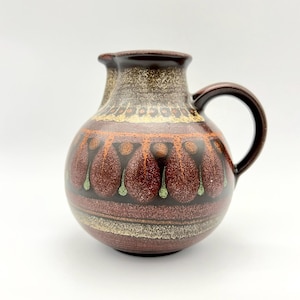 May include: A brown ceramic pitcher with a geometric design in shades of orange, green, and white. The pitcher has a rounded body and a handle.