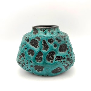 May include: A squat, rounded ceramic vase featuring a heavily textured, porous surface with a mottled pattern of deep black and vibrant turquoise glaze. The vessel has a wide, bulbous body that tapers slightly towards a smooth, dark rim.