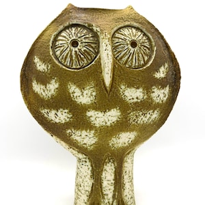 May include: A ceramic owl figurine featuring a textured brown and cream glaze. The owl has large, circular, deeply incised eyes, a pointed beak, and a wide, rounded head that tapers down into a narrow, cylindrical base with vertical markings.