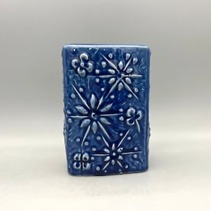Puede incluir: Un pequeño jarrón de cerámica azul con forma cuadrada y un diseño floral en relieve. El jarrón tiene una superficie texturizada y un acabado brillante.