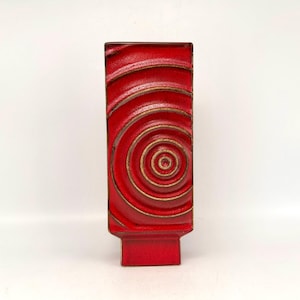 Könnte beinhalten: Eine rechteckige rote Keramikvase mit einem Spiraldesign. Die Vase hat eine strukturierte Oberfläche mit konzentrischen Kreisen in einem helleren Rotton und goldenen Akzenten. Der Sockel ist ein durchgehend rotes Rechteck.