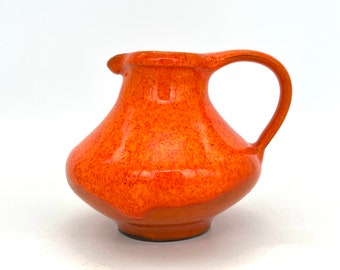 May include: A small orange ceramic pitcher with a speckled glaze. The pitcher has a rounded body and a short, curved spout.