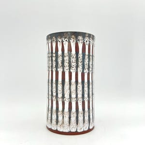 Puede incluir: Un jarrón cilíndrico de cerámica con superficie texturizada. El jarrón presenta un patrón de rayas verticales en blanco, gris oscuro y marrón rojizo. El jarrón mide unos 25 cm de alto.