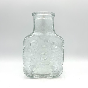 Könnte beinhalten: Transparente Glasflasche mit strukturierter Oberfläche und einer einzigartigen, abgerundeten rechteckigen Form. Die Flasche hat sechs kreisförmige Vertiefungen und eine strukturierte Oberfläche. Die Flasche ist leer und steht vor einem weißen Hintergrund.