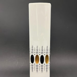 Puede incluir: Un jarrón blanco, rectangular y alto, con un patrón decorativo. El patrón incluye óvalos negros, dorados y negros, junto con pequeños círculos y espirales negros. El jarrón tiene un acabado brillante y está sobre un fondo oscuro.