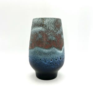 May include: A ceramic vase with a unique, textured surface. The vase features a gradient of colors, including dark blue at the base, transitioning to teal, then a band of reddish-brown, and finally a speckled gray at the top. The vase has a rounded shape.