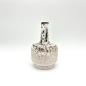 May include: A small, white ceramic vase with a brown speckled pattern. The vase has a round base and a narrow neck.