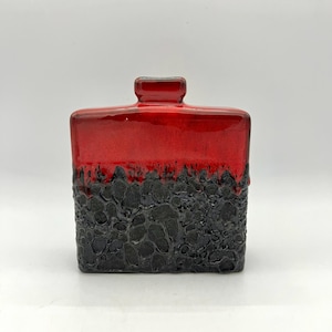 May include: A square, ceramic vase with a glossy red top and a textured, black bottom. The vase has a rectangular opening and a unique, lava-like texture on the lower half. The vase is a decorative item.