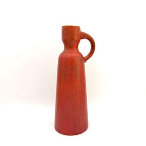 Può includere: Un vaso in ceramica rosso con manico. Il vaso ha una superficie strutturata e un collo stretto che si allarga verso la base. Il manico è un anello arrotondato. Il vaso è di colore rosso pieno con qualche macchiolina più scura.