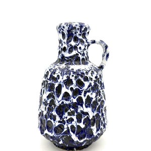 May include: A ceramic pitcher featuring a textured, cratered surface with a high-contrast glaze pattern. The vessel is finished in deep navy blue and bright white, with a thick, dripping, marbled effect covering the entire exterior and handle.