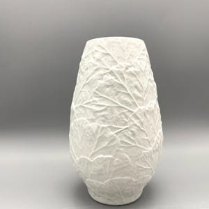 Puede incluir: Jarrón de cerámica blanca con un patrón de hojas texturizadas. El jarrón tiene una base ancha que se estrecha hacia arriba, con una abertura ligeramente ensanchada. La superficie está cubierta con un diseño detallado de hojas en relieve.