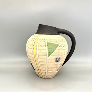 May include: A ceramic jug with a black handle and a geometric design in shades of yellow, green, blue, and pink. The jug has a white background with a woodgrain texture.