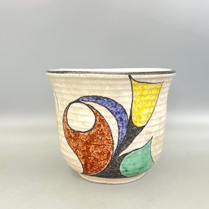 Puede incluir: Un macetero de cerámica blanca con un diseño floral estilizado en amarillo, azul, verde y marrón. El macetero tiene una superficie texturizada y un borde ligeramente acampanado.