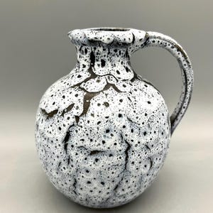 May include: A white ceramic pitcher with a speckled glaze and a brown handle. The pitcher has a round body and a narrow neck.
