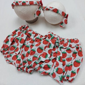 K&ouml;nnte beinhalten: Ein Baby-Outfit-Set mit einem wei&szlig;en Hintergrund und einem roten Erdbeermuster. Das Set enth&auml;lt einen Bloomer und zwei passende Stirnb&auml;nder. Die Stirnb&auml;nder haben eine kleine rote Schleife. Der Bloomer hat einen ger&uuml;schten Bund und Bein&ouml;ffnungen.