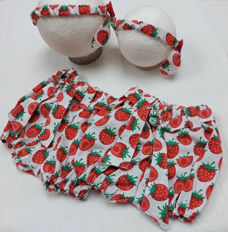 K&ouml;nnte beinhalten: Ein Baby-Outfit-Set mit einem wei&szlig;en Hintergrund und einem roten Erdbeermuster. Das Set enth&auml;lt einen Bloomer und zwei passende Stirnb&auml;nder. Die Stirnb&auml;nder haben eine kleine rote Schleife. Der Bloomer hat einen ger&uuml;schten Bund und Bein&ouml;ffnungen.