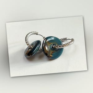 May include: A pair of silver hoop earrings with teal and gold glass disc charms. The charms have a swirling pattern.