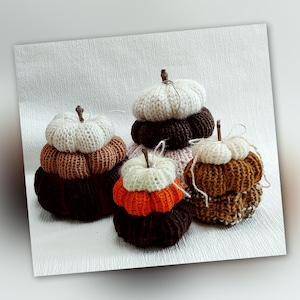 Op de afbeelding: Vier gebreide pompoenen in verschillende tinten bruin, oranje en wit. De pompoenen zijn op elkaar gestapeld, waardoor een gelaagd effect ontstaat.