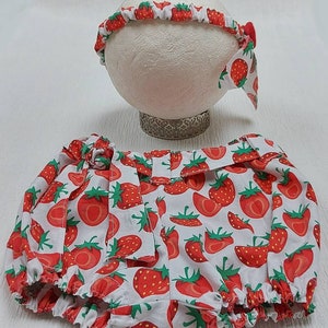 K&ouml;nnte beinhalten: Ein Baby-Outfit-Set mit einem Stirnband und Bloomers, beide mit einem leuchtenden Erdbeermuster auf wei&szlig;em Hintergrund verziert. Das Stirnband hat eine kleine Schleife, und die Bloomers haben eine passende Schleife an der Taille. Das Set ist f&uuml;r Babys konzipiert.