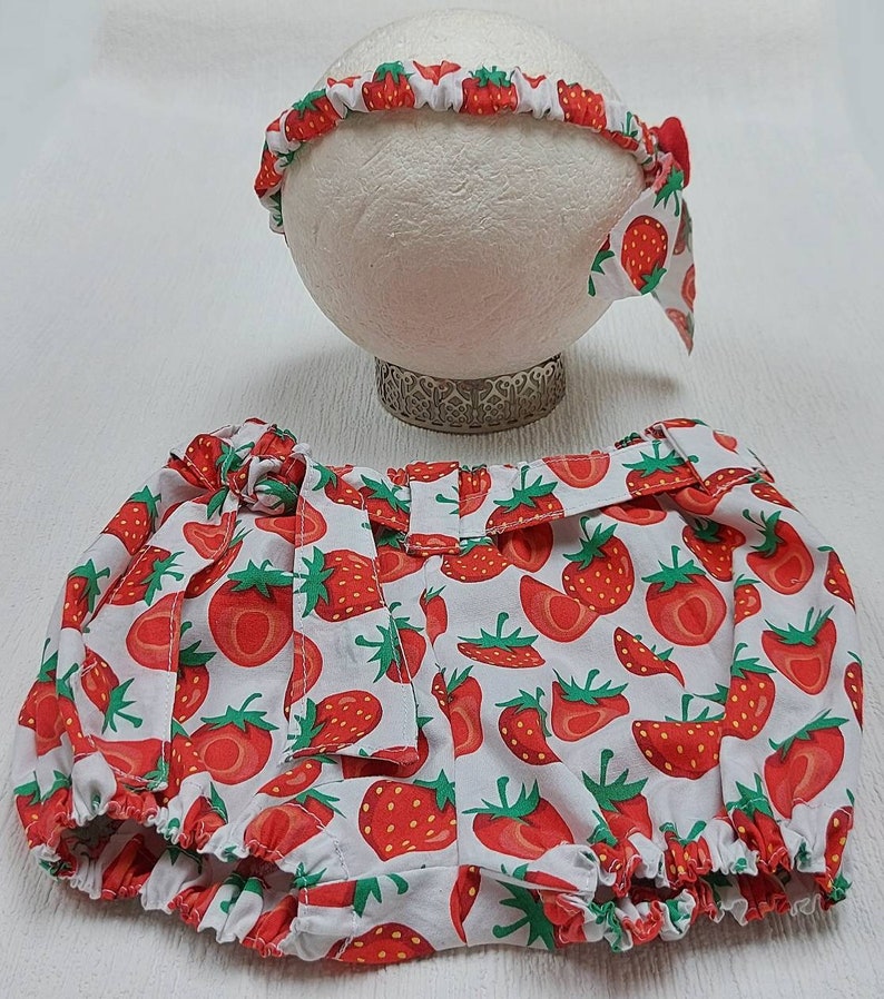K&ouml;nnte beinhalten: Ein Baby-Outfit-Set mit einem Stirnband und Bloomers, beide mit einem leuchtenden Erdbeermuster auf wei&szlig;em Hintergrund verziert. Das Stirnband hat eine kleine Schleife, und die Bloomers haben eine passende Schleife an der Taille. Das Set ist f&uuml;r Babys konzipiert.