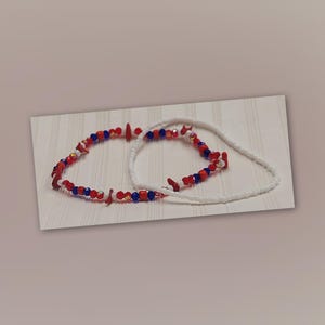 Op de afbeelding: Twee armbanden met kralen. Eén armband heeft rode, witte en blauwe kralen, de andere is wit. De armbanden zijn gerangschikt op een wit oppervlak met een subtiel streepjespatroon. De kralen zijn klein en rond.