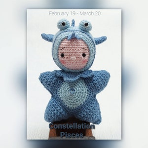 May include: A crocheted plush toy in the shape of a fish wearing a blue and light blue crocheted costume with a star shape. The toy has a pink face with black eyes and a small pink nose. The text "Constellation Pisces" is written below the toy.