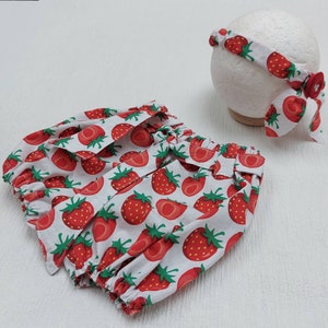 K&ouml;nnte beinhalten: Ein wei&szlig;es Baby-Outfit mit einem roten Erdbeermuster. Das Set beinhaltet eine Pumphose und ein passendes Stirnband. Das Stirnband ist mit einer roten Schleife und einem dekorativen Knopf verziert.