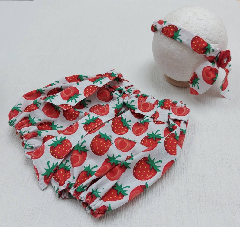 K&ouml;nnte beinhalten: Ein wei&szlig;es Baby-Outfit mit einem roten Erdbeermuster. Das Set beinhaltet eine Pumphose und ein passendes Stirnband. Das Stirnband ist mit einer roten Schleife und einem dekorativen Knopf verziert.