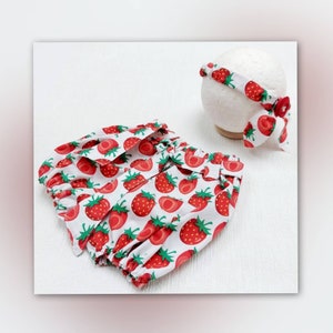 K&ouml;nnte beinhalten: Ein Baby-Outfit-Set mit einer Pumphose und einem Stirnband, beide mit einem leuchtend roten Erdbeermuster auf wei&szlig;em Hintergrund verziert. Das Stirnband enth&auml;lt eine dekorative Schleife mit passendem Erdbeermuster.