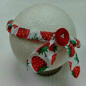 K&ouml;nnte beinhalten: Ein Stoff-Haarband mit einem sich wiederholenden Muster aus roten Erdbeeren auf wei&szlig;em Grund. Das Haarband hat eine rote Stoffblume mit drei kleinen, silbernen Perlen. Das Haarband ist um eine wei&szlig;e, kugelf&ouml;rmige Form gewickelt.