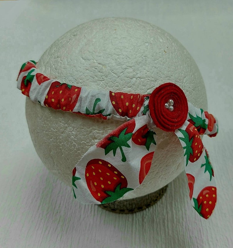 K&ouml;nnte beinhalten: Ein Stoff-Haarband mit einem sich wiederholenden Muster aus roten Erdbeeren auf wei&szlig;em Grund. Das Haarband hat eine rote Stoffblume mit drei kleinen, silbernen Perlen. Das Haarband ist um eine wei&szlig;e, kugelf&ouml;rmige Form gewickelt.