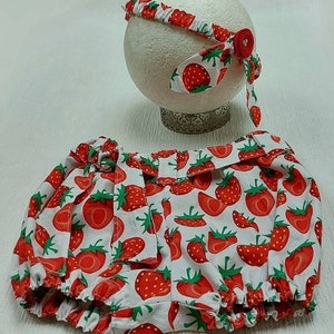 K&ouml;nnte beinhalten: Ein Baby-Outfit-Set mit Stirnband und Bloomers. Beide Artikel sind wei&szlig; mit einem sich wiederholenden Muster aus roten Erdbeeren und gr&uuml;nen Bl&auml;ttern. Das Stirnband hat einen roten Blumenakzent. Die Bloomers haben einen ger&uuml;schten Rand und eine Schleife.