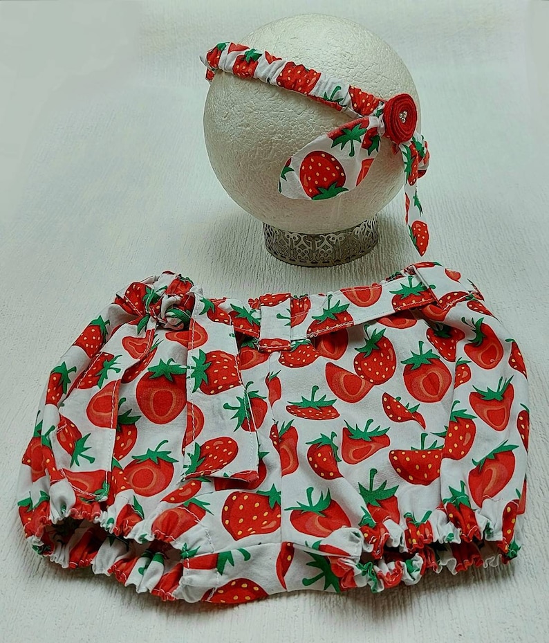 K&ouml;nnte beinhalten: Ein Baby-Outfit-Set mit Stirnband und Bloomers. Beide Artikel sind wei&szlig; mit einem sich wiederholenden Muster aus roten Erdbeeren und gr&uuml;nen Bl&auml;ttern. Das Stirnband hat einen roten Blumenakzent. Die Bloomers haben einen ger&uuml;schten Rand und eine Schleife.