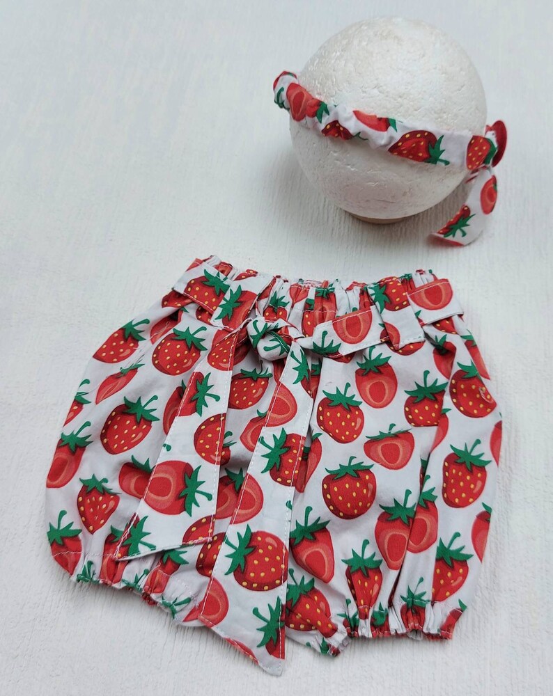 K&ouml;nnte beinhalten: Ein Baby-Outfit mit wei&szlig;en Shorts und einem passenden Stirnband, beide mit einem leuchtend roten Erdbeermuster verziert. Die Shorts haben eine Schleife an der Taille. Das Stirnband ist um eine wei&szlig;e Kugel gelegt.
