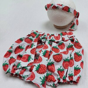 K&ouml;nnte beinhalten: Ein Baby-Outfit-Set mit wei&szlig;em Hintergrund und einem roten Erdbeermuster. Das Set beinhaltet einen Bloomer mit Schleifen und ein passendes Stirnband. Das Stirnband wird auf einer wei&szlig;en Kugelform pr&auml;sentiert.