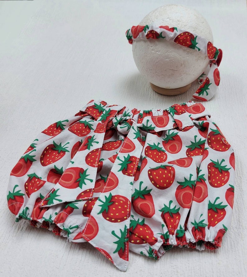 K&ouml;nnte beinhalten: Ein Baby-Outfit-Set mit wei&szlig;em Hintergrund und einem roten Erdbeermuster. Das Set beinhaltet einen Bloomer mit Schleifen und ein passendes Stirnband. Das Stirnband wird auf einer wei&szlig;en Kugelform pr&auml;sentiert.