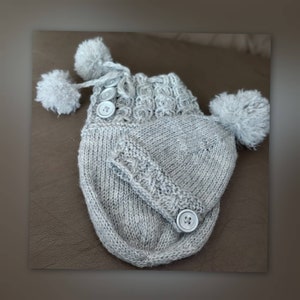 May include: Gray knitted baby hat with a matching gray knitted mitten attached. The hat has a pom-pom on top and the mitten has a button.