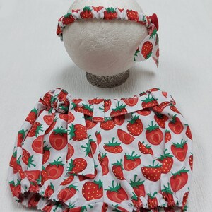 K&ouml;nnte beinhalten: Ein Baby-Outfit-Set mit Stirnband und Bloomers. Beide Artikel sind wei&szlig; mit einem sich wiederholenden Muster aus roten Erdbeeren und gr&uuml;nen Bl&auml;ttern. Das Stirnband hat eine kleine rote Schleife. Die Bloomers haben einen ger&uuml;schten Bund und Bein&ouml;ffnungen.