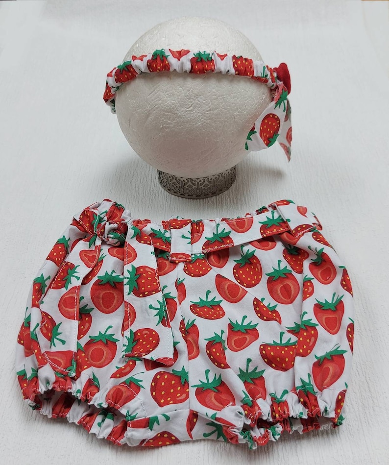K&ouml;nnte beinhalten: Ein Baby-Outfit-Set mit Stirnband und Bloomers. Beide Artikel sind wei&szlig; mit einem sich wiederholenden Muster aus roten Erdbeeren und gr&uuml;nen Bl&auml;ttern. Das Stirnband hat eine kleine rote Schleife. Die Bloomers haben einen ger&uuml;schten Bund und Bein&ouml;ffnungen.