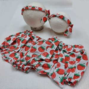 K&ouml;nnte beinhalten: Ein Set Babykleidung mit Erdbeermuster. Das Set enth&auml;lt eine Pumphose und zwei Stirnb&auml;nder. Die Pumphose und die Stirnb&auml;nder sind wei&szlig; mit einem sich wiederholenden Muster aus roten Erdbeeren und gr&uuml;nen Bl&auml;ttern. Die Stirnb&auml;nder haben eine rote Schleife.