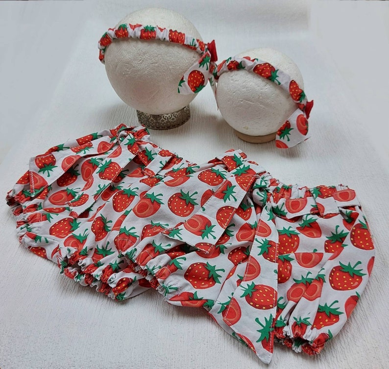 K&ouml;nnte beinhalten: Ein Set Babykleidung mit Erdbeermuster. Das Set enth&auml;lt eine Pumphose und zwei Stirnb&auml;nder. Die Pumphose und die Stirnb&auml;nder sind wei&szlig; mit einem sich wiederholenden Muster aus roten Erdbeeren und gr&uuml;nen Bl&auml;ttern. Die Stirnb&auml;nder haben eine rote Schleife.