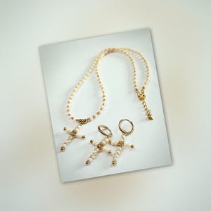 Op de afbeelding: Een crèmekleurige ketting en bijpassende oorbellen set. De ketting en oorbellen hebben goudkleurige kruisvormige hangers. De oorbellen zijn van het type oorring. De sieraden worden op een wit oppervlak gepresenteerd.