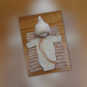 Puede incluir: Un conjunto de bebé de punto blanco con un gorro a juego, colocado sobre una manta de rayas marrones y blancas.