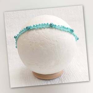 Op de afbeelding: Een lichtblauw hoofdband met een gehaakt kantontwerp en turquoise kralen.